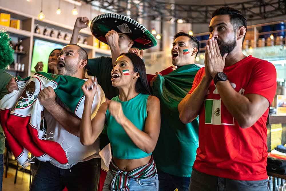 Grupo de aficionados mexicanos celebrando y reaccionando durante un partido mientras ven la copa del mundo juntos en un bar con colores de la selección.